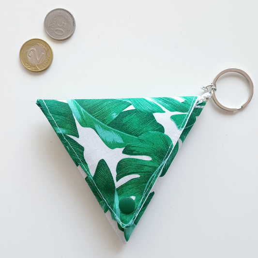 Μίνι πορτοφολάκι origami σε﻿ λευκό ύφασμα με σχέδιo﻿  πράσινα φύλλα monstera.﻿ Κούμπωμα πράσινο και﻿ αλυσίδα με μπρελόκ. Από πάνω υπάρχουν δυο νομίσματα.