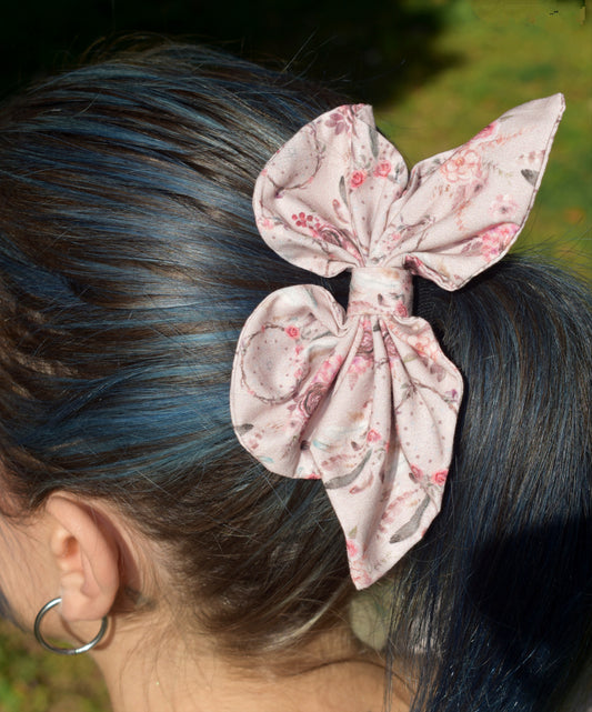 Υφασμάτινη πεταλούδα σε απαλό ροζ χρώμα με σχεδια λουλουδάκια και ονειροπαγίδες σε αντίστοιχες αποχρώσεις. Είναι φορεμένη σε μαλλιά καστανά με μπλε ανταύγειες. 
