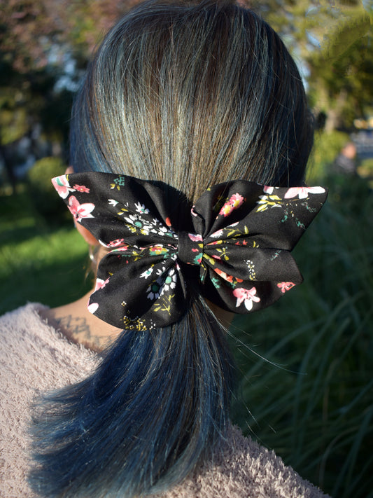 Υφασμάτινη μαύρη πεταλούδα με λουλούδια διαφόρων χρωμάτων και μεγεθών. Είναι φορεμένη σε καστανά μαλλιά με μπλε ανταύγειες.