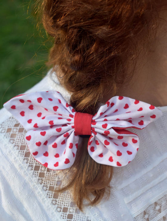 Υφασμάτινη λευκή πεταλούδα με κοκκινες καρδούλες και κόκκινη λωρίδα στη μέση. Είναι φορεμένη σε καστανοκόκκινα μαλλιά.